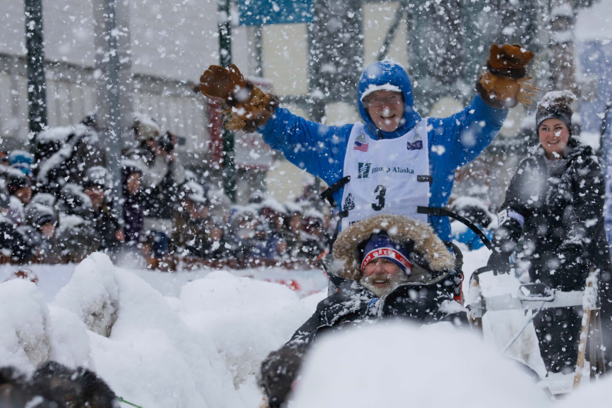 A man with a light blue parka holds his arms up as he stands on a sled in heavy snow. A nother man in a fur-ruffed parka sits on the sled in front of him and crowds watch on the sidelines.