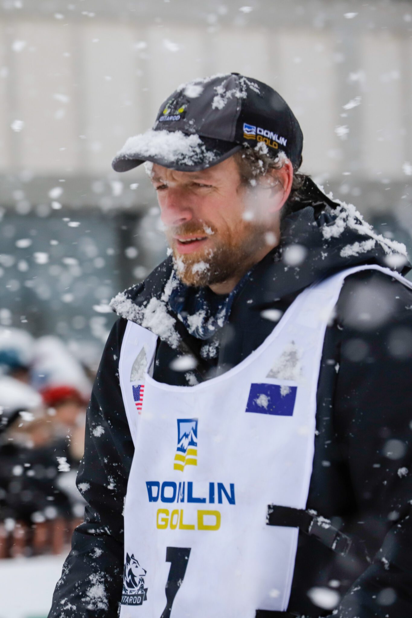 A man with reddish hair and wearing a thick black parka and a white bib stares ahead in heavy snow.