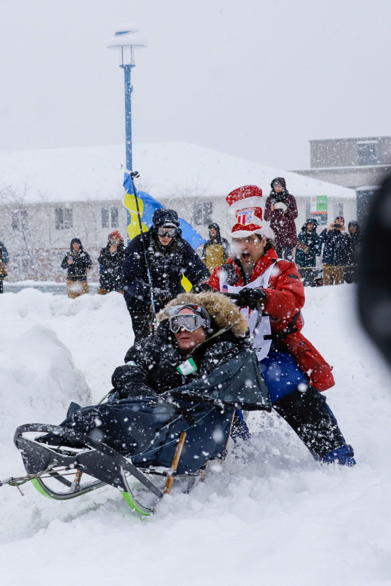 three people in a dog sled caravan: a person in the sled, a person in a Dr. Seuss hat, and another with the Ukrainian flag