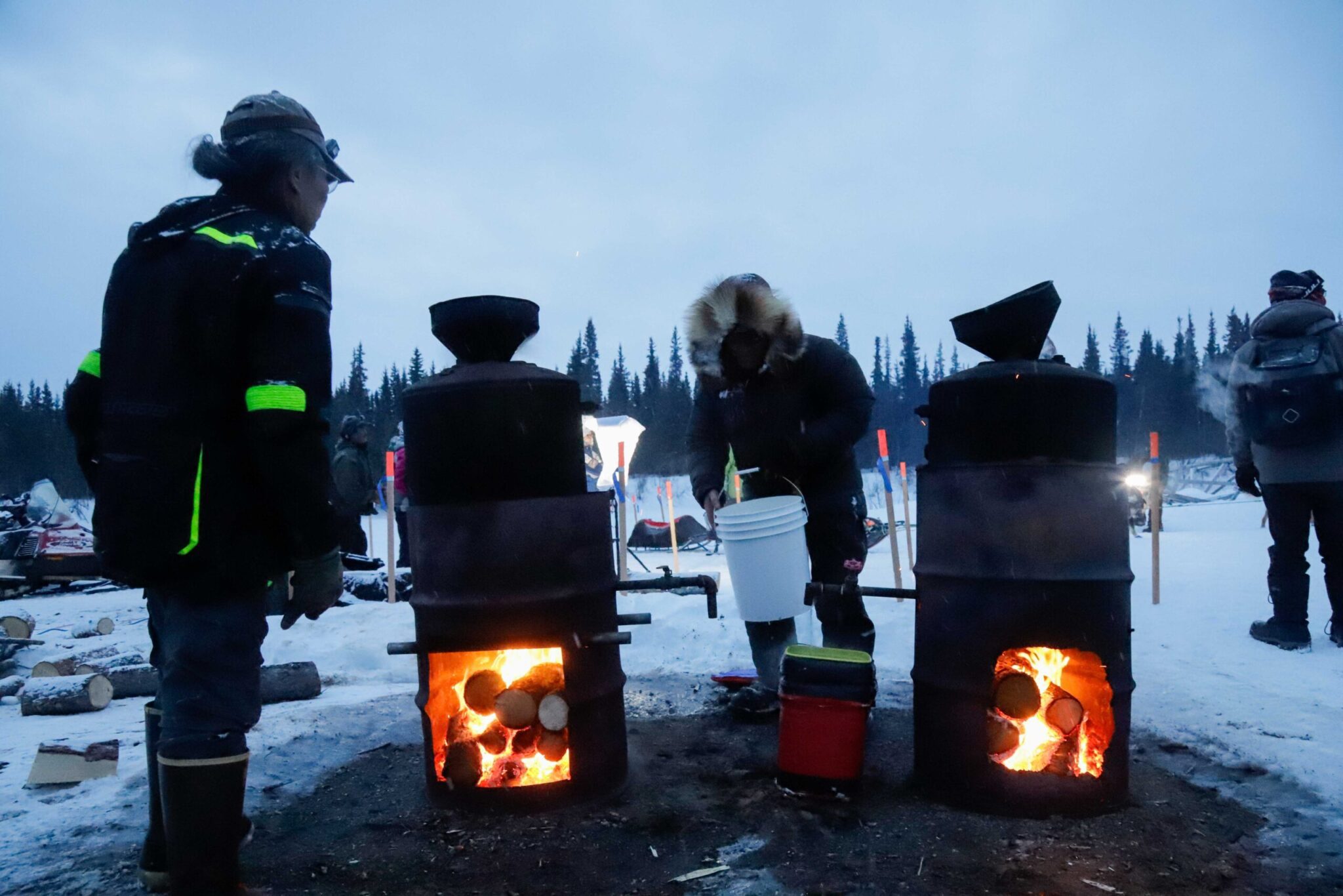 two people stand near two burn barrels that are heating up water