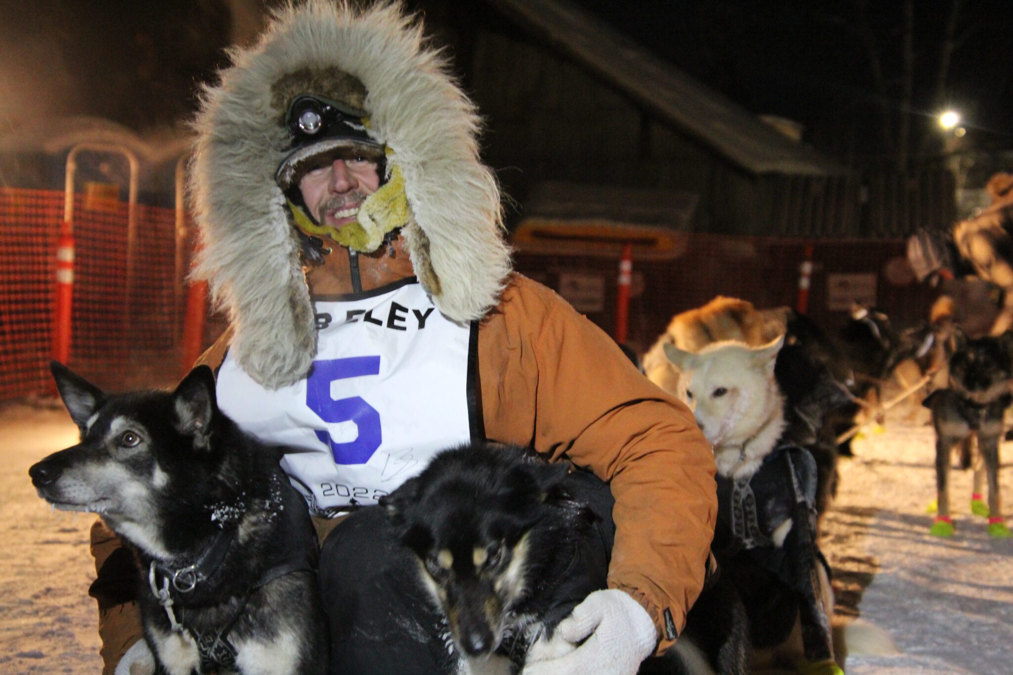 A man in an orange parka smiles for the camera while holding his two dogs