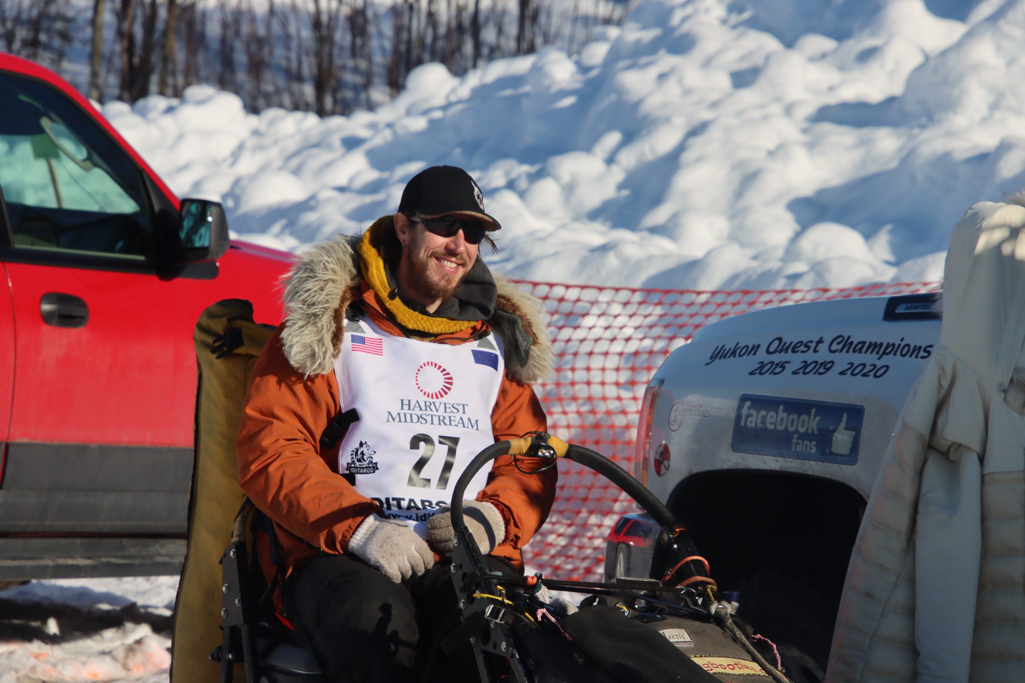 A man in an orange parka sits on a sled next to a truck