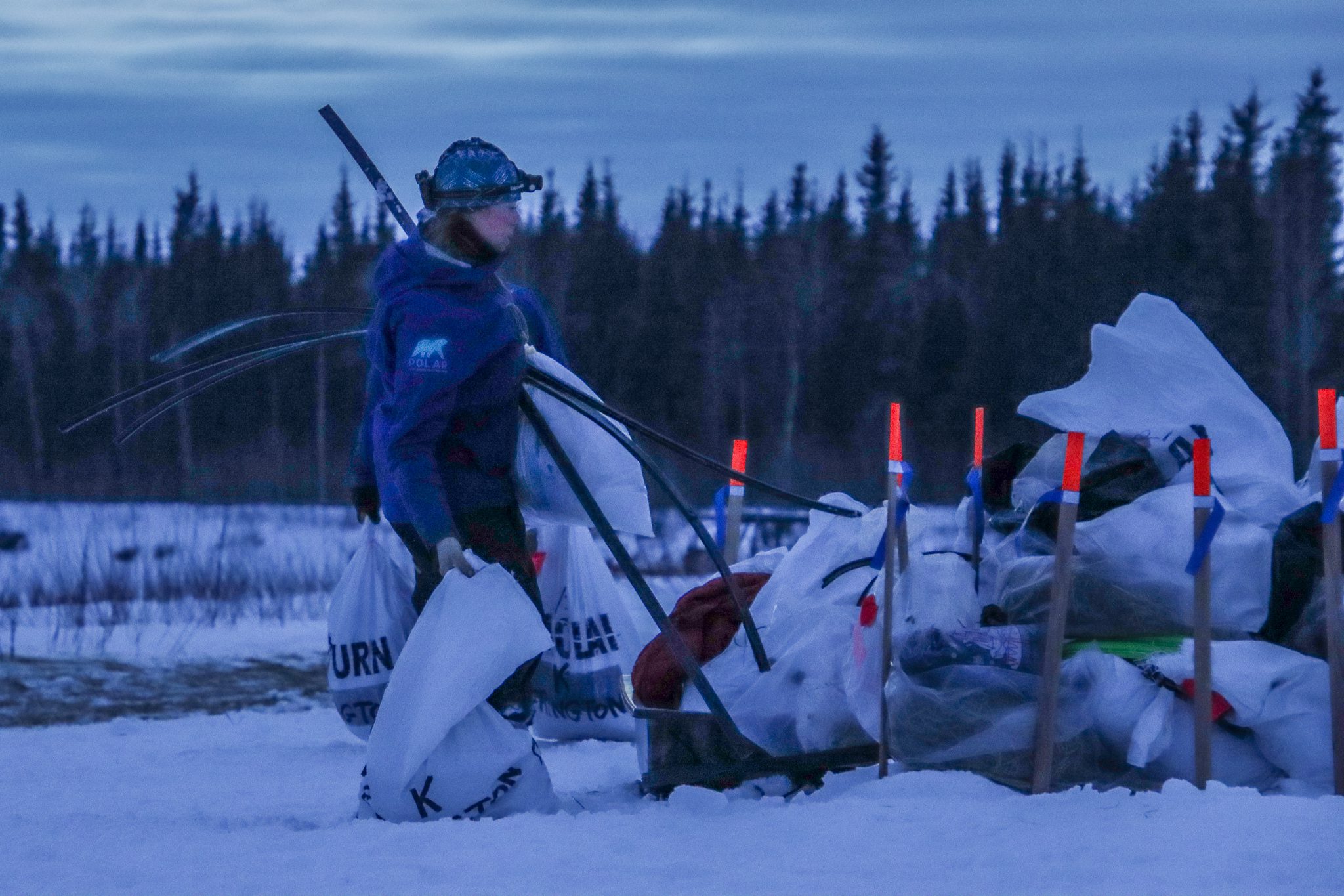 A person carrying white canvas bags and black strips of plastic in a snowy area