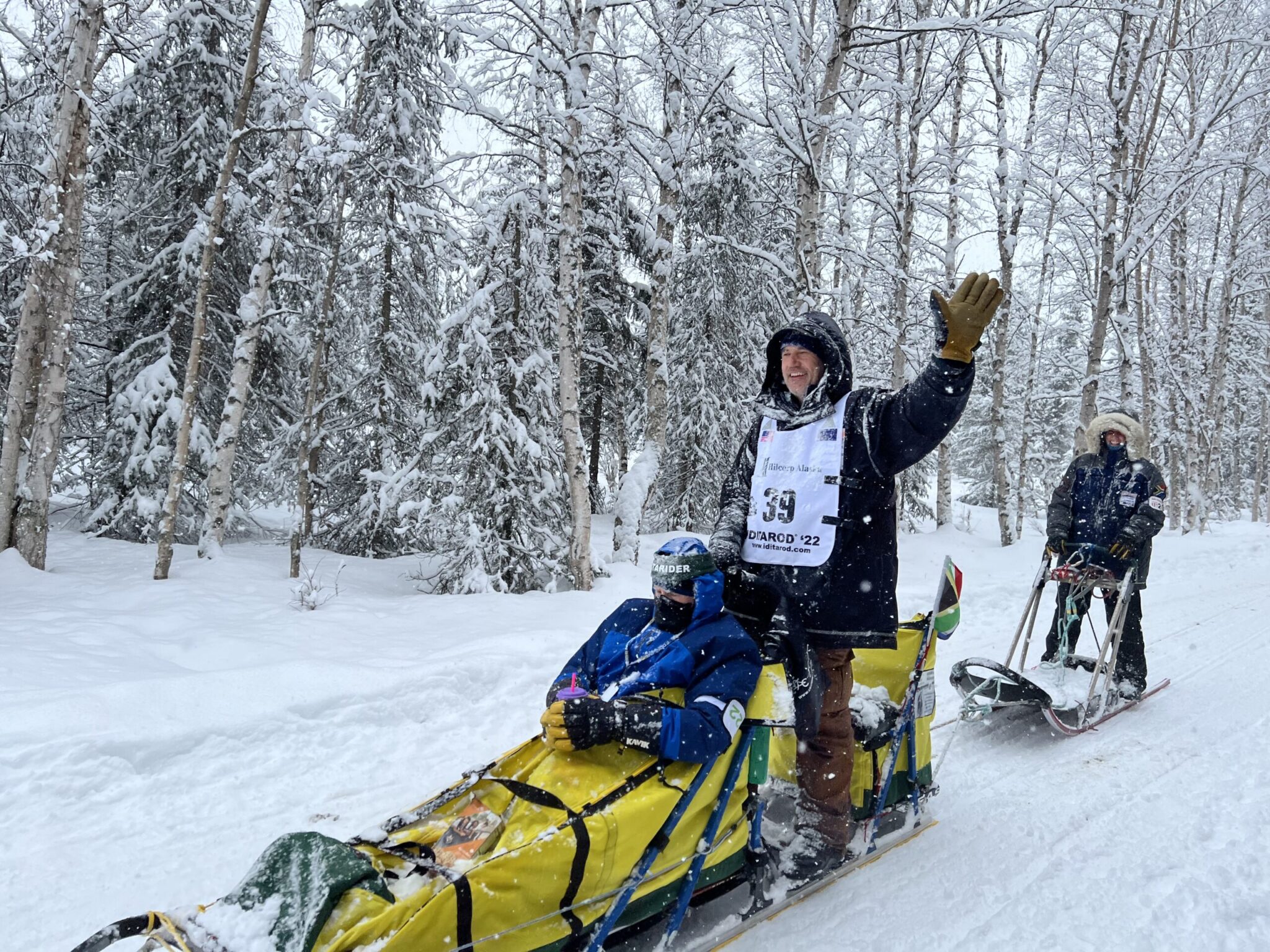 a musher waves from the back of a sled on a snowy day