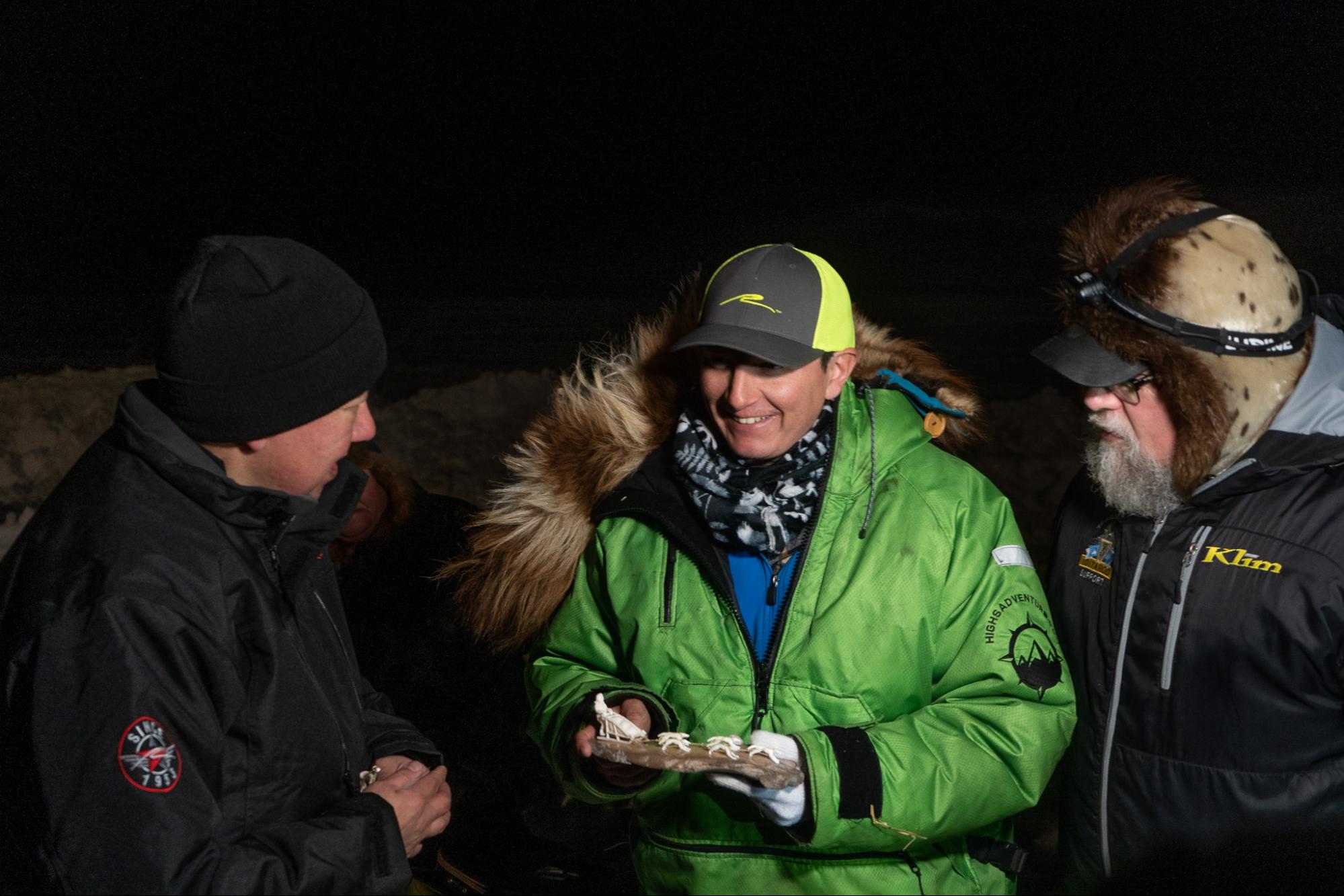 a musher in a green jacket holds a ivory carving