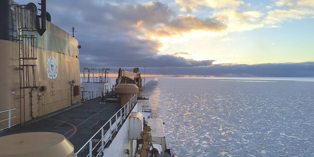 An image showing the side of the U.S. Coast Guard Cutter Healy looking towards her stern as she break a path through icy waters with the sun setting in the background. Photo courtesy of NOAA. 