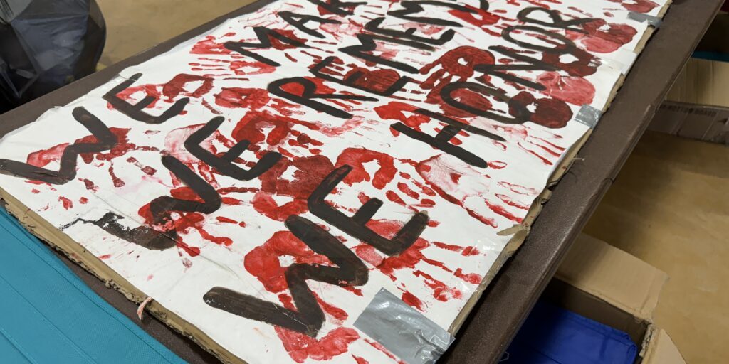 A sign with red paint that says "WE MARCH, WE REMEMBER, WE HONOR" at the Village of Solomon's 2024 Missing and Murdered Indigenous Persons Day event. Ben Townsend photo. 