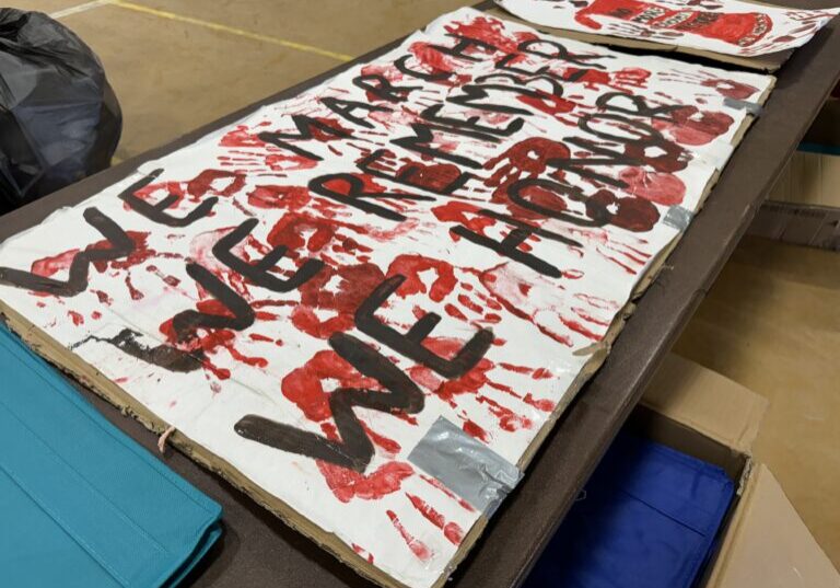 A sign with red paint that says "WE MARCH, WE REMEMBER, WE HONOR" at the Village of Solomon's 2024 Missing and Murdered Indigenous Persons Day event. Ben Townsend photo. 