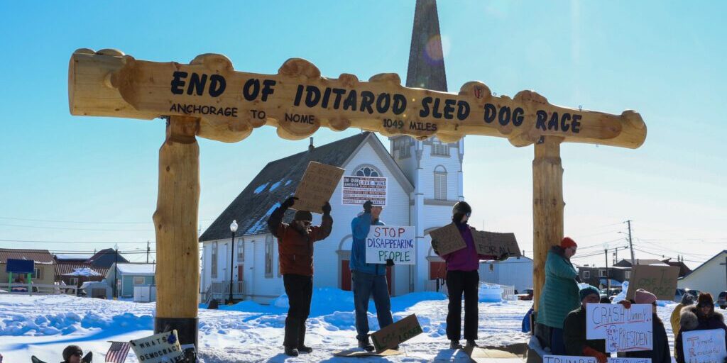 Protestors hold signs on the base of the Iditarod's burled arch in Anvil City Square. Wali Rana photo. 
