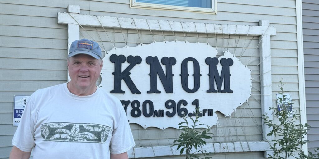 Former KNOM Radio Engineer John Pfeifer poses for a photo in front of a KNOM Radio sign. Sarah Swartz photo.